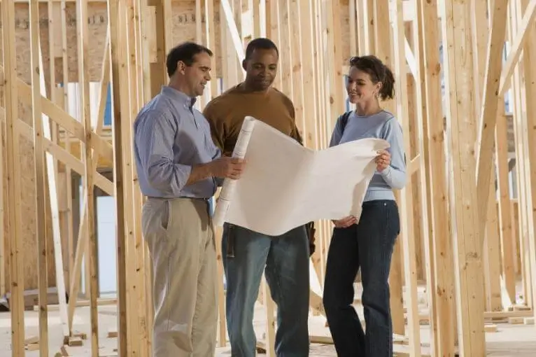 Group of three people, including a couple and a contractor, reviewing blueprints at a construction site with exposed wooden framing, discussing home building plans and design.